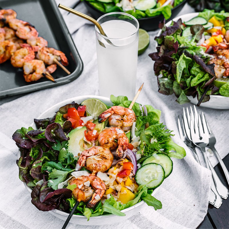Salad with a skewer of shrimp laid across it, salad set in bowl on a table cloth on a table outdoors with other salads beside it