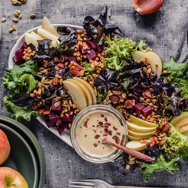 Roasted beet and apple salad on a glass plate on a table cloth with a bowl of apples on it