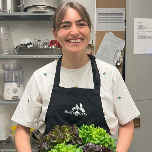 Woman smiling in a kitchen wearing an apron and holding a bunch of leafy greens